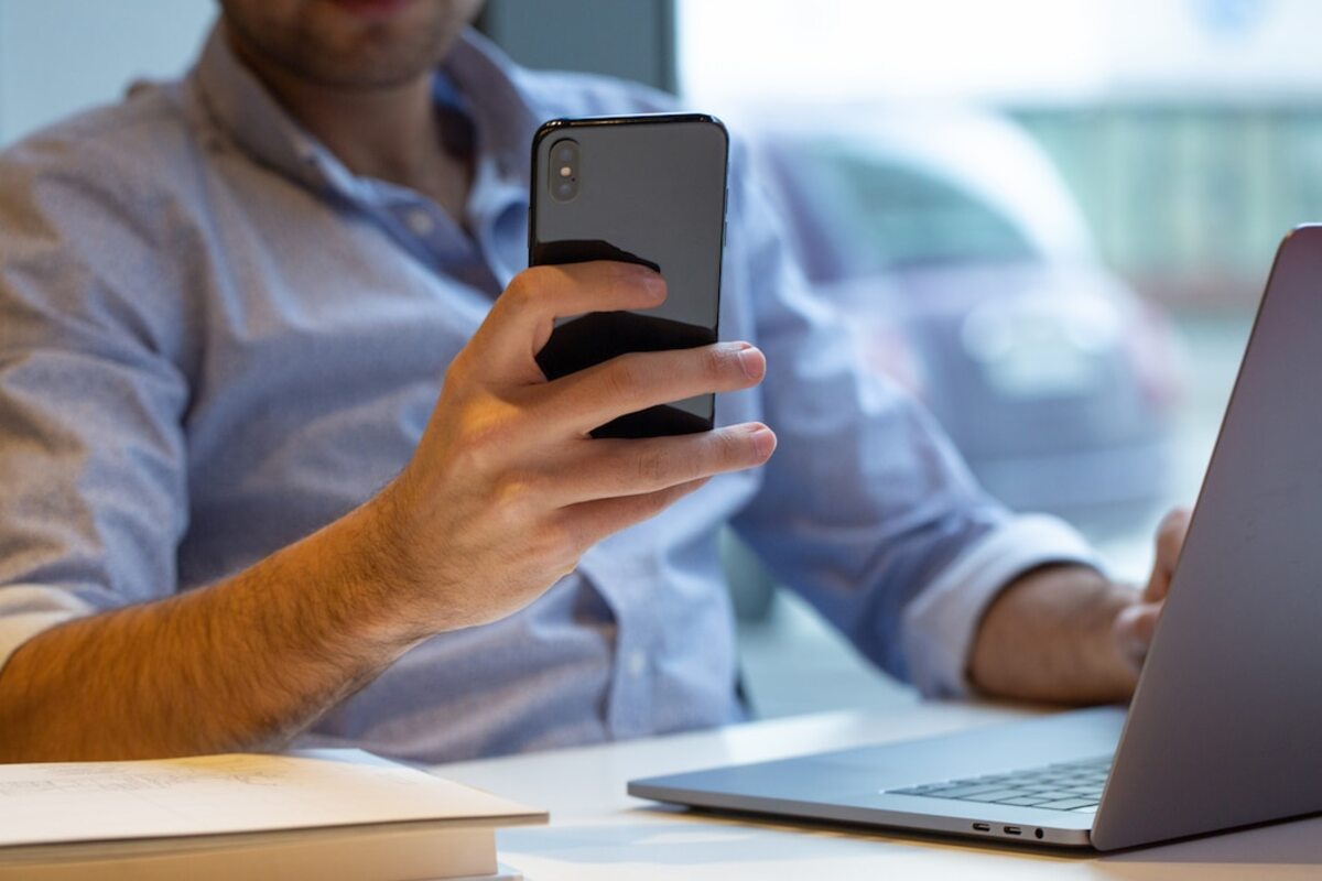 homem com blusa social azul sentado em uma mesa com um laptop na sua frente. Eles está segurando um celular