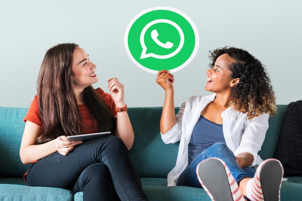 duas mulheres sentadas em um sofá. Uma delas está com um tablet na mão enquanto a outra, segura o símbolo do WhatsaApp