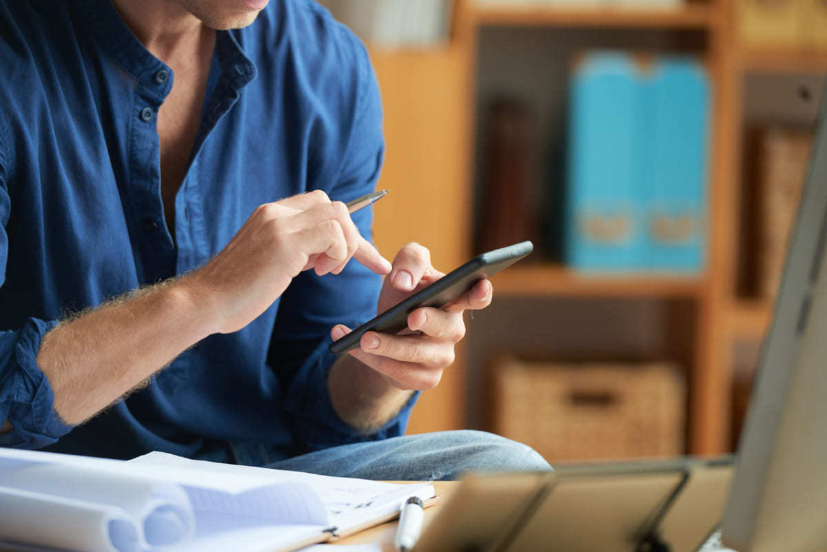 homem vestindo camisa social azul, sentado com celular na mão e relatórios espalhados em cima da mesa