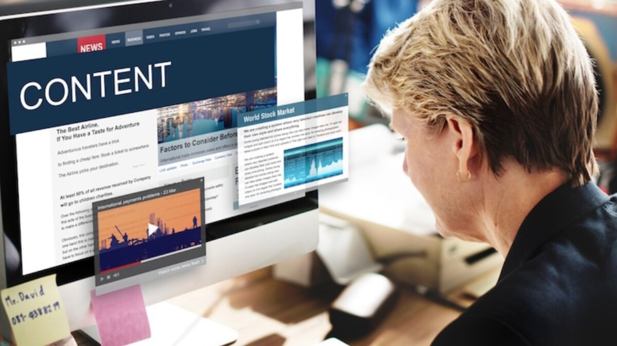 mulher loira de cabelo curto sentada em frente a um monitor com uma matéria jornalística sendo exposta na tela dele