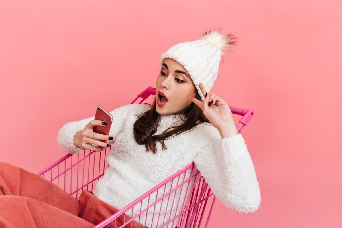 Uma mulher vestindo uma touca e blusa branca está sentada em um carrinho de supermercado cor de rosa, em um cenário rosa. Ela segura um celular na mão enquanto tira uma selfie.