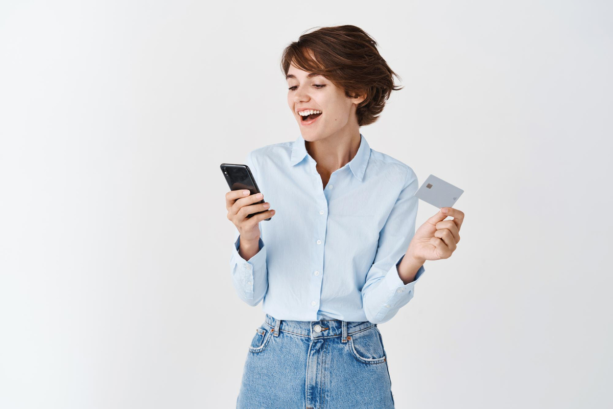 mulher com bulsa social e calça jeans com expressão alegre, segurando um celular em uma mão e, em outra, um cartão