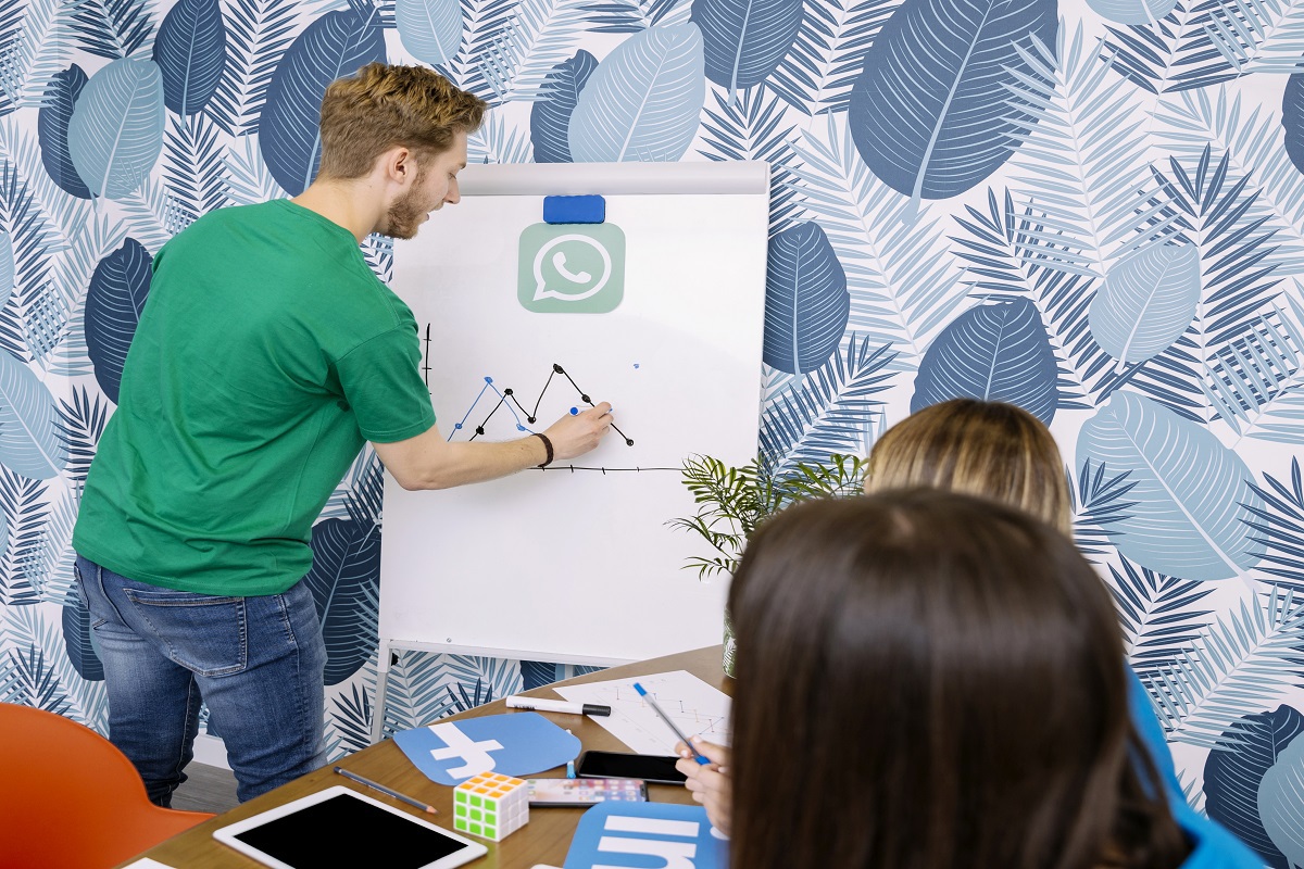 homem de pé, desenhando um gráfico de linhas em um quadro branco. Ele parece apresentar uma estratégia para as outras pessoas que estão na foto