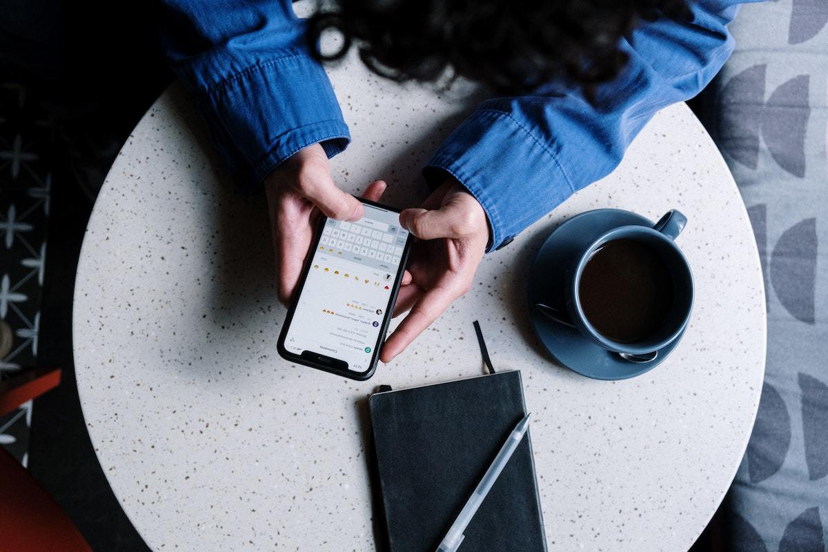 pessoa sentada, usando o celular. Na mesa também há um caderno, uma caneta e uma caneca de café