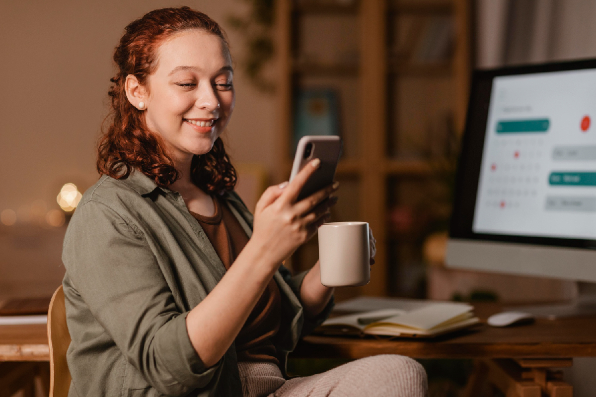 mulher sorrindo, sentada, com uma caneca de café e olhando o celular