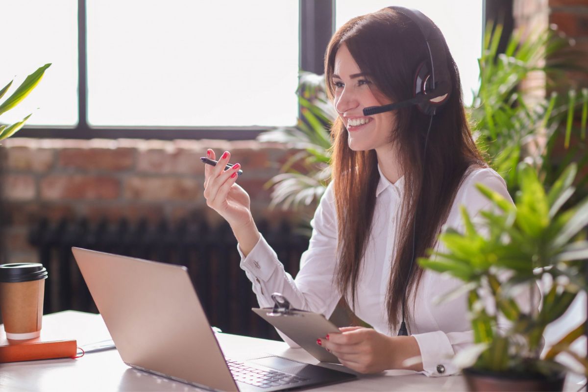 Mulher sorridente sentada diante de mesa usando notebook e fones de ouvido enquanto aparentemente realiza um atendimento online