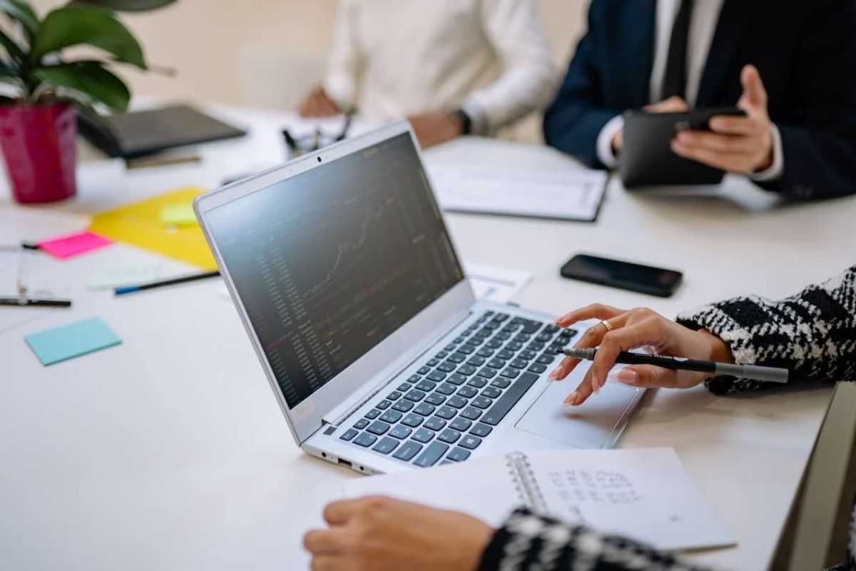 Pessoas ao redor de mesa branca de reunião com uma delas usando notebook e alguns objetos como post-its espalhados ao redor dos membros da reunião