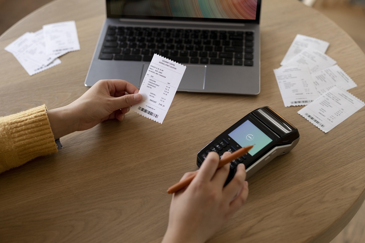 Mulher diante de mesa de madeira com maquineta de cartão de crédito e um ticket de pagamento na mão esquerda, enquanto segura lápis, além de notebook ao fundo da imagem com outros tickets espalhados