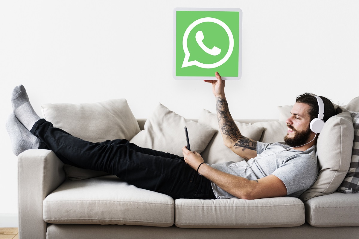 Homem deitado em sofá usando fones de ouvido e segurando smartphone com mão esquerda e placa com símbolo do whatsapp com a direita