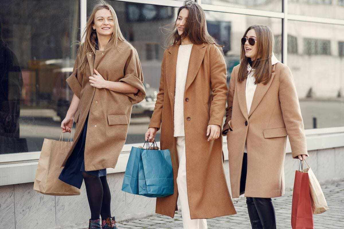 Três mulheres andando lado a lado em calçada de avenida diante de vidraça de prédio enquanto sorriem e carregam sacolas de compras de várias cores e tamanhos