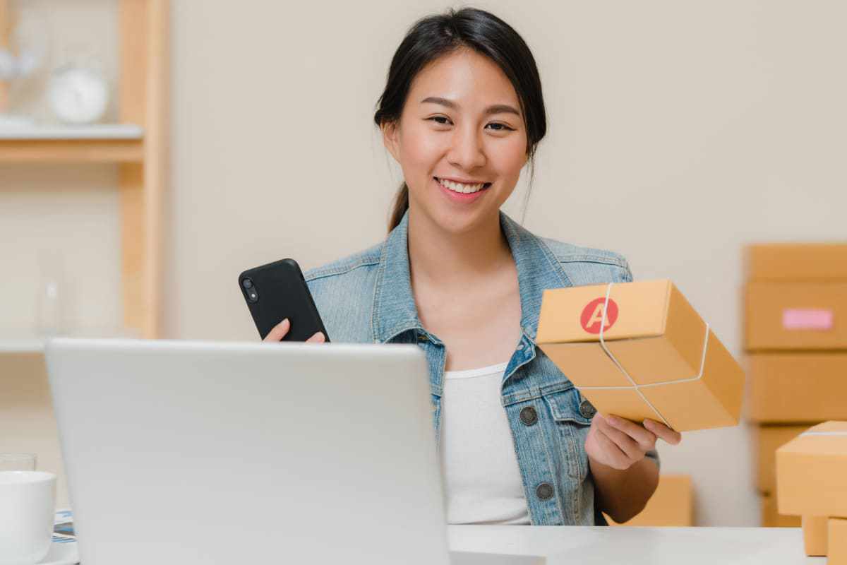 Mulher diante de notebook com caixa de entrega na mão e celular na outra despachando mercadoria que teve saída com métodos de Como fazer vendas online