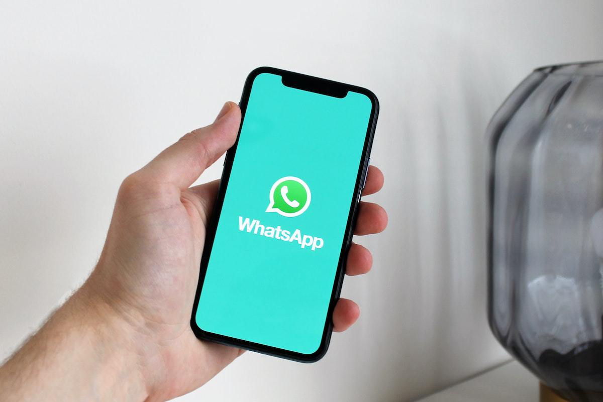 Pessoa segurando celular com mão esquerda no interior de local com ambiente branco em desfoco ao fundo, com detalhes na cor cinza, enquanto o aparelho exibe tela de inicialização do aplicativo WhatsApp, local onde é possível fazer boas Vendas como afiliado