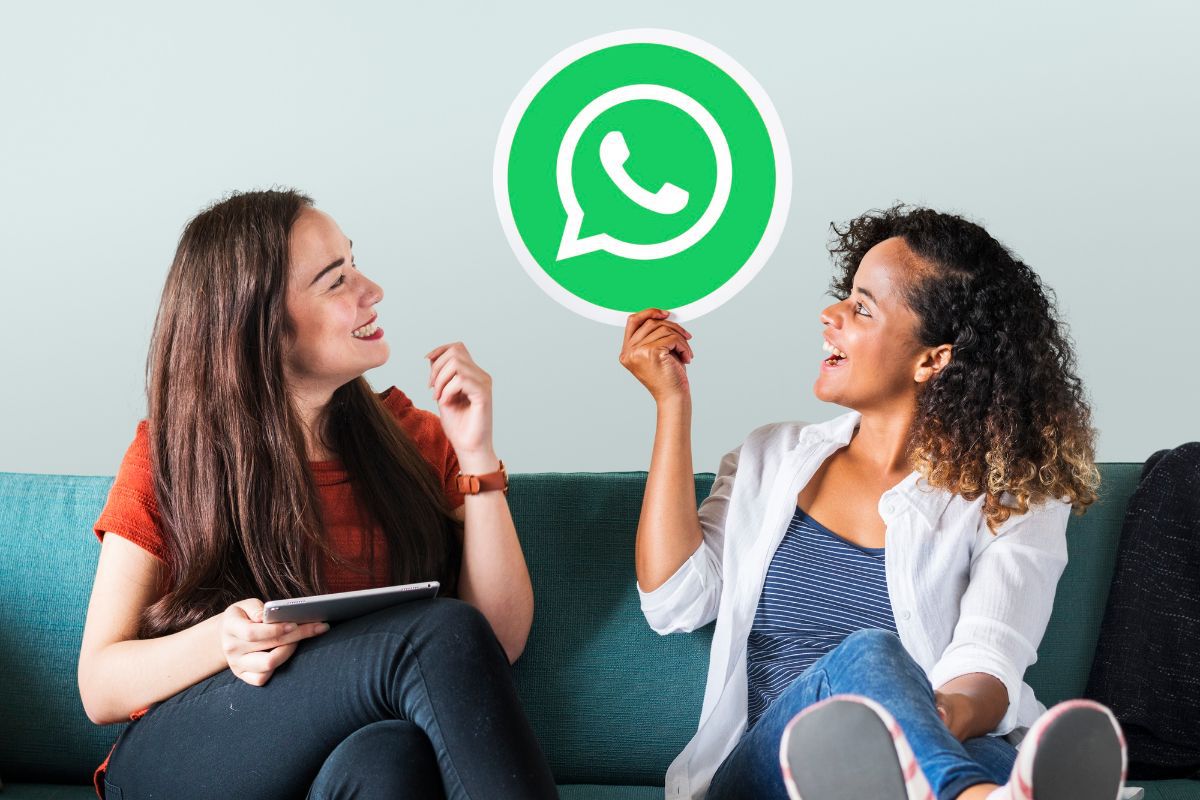 Duas mulheres sentadas lado a lado em sofá se olham e sorriem enquanto conversam e uma delas segura placa com símbolo do app do whatsapp, onde é possível realizar Vendas consultivas