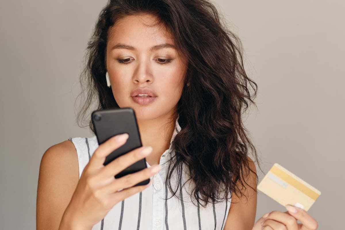Mulher segurando celular com uma mão e cartão de crédito com a outra com semblante sério e mostrando concentração ao fazer compra de empresa que realiza Vendas corporativas pelo WhatsApp